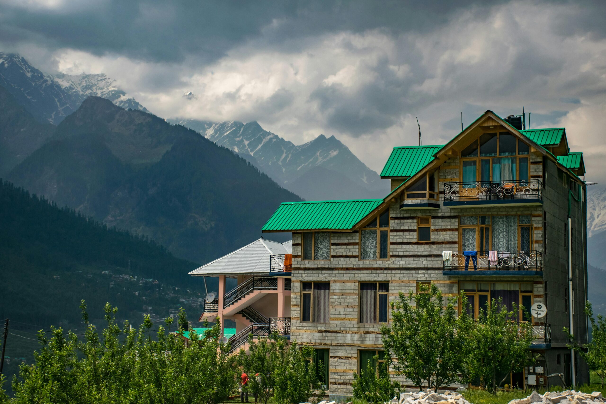 Modern mountain house with green roof, set against snow-capped peaks under cloudy skies.